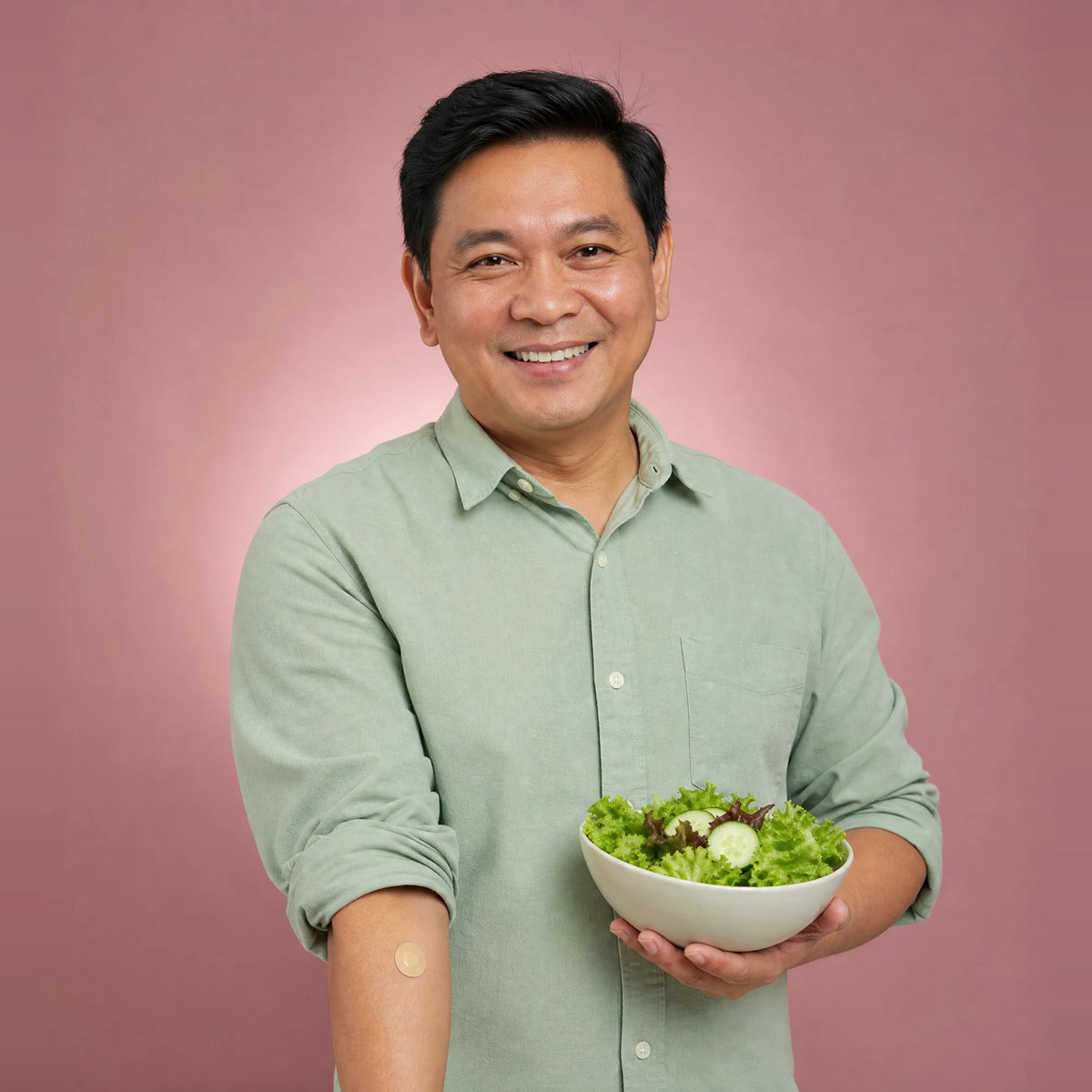 Smiling man holding a bowl of green salad, with a small plaster on his arm after a home blood test